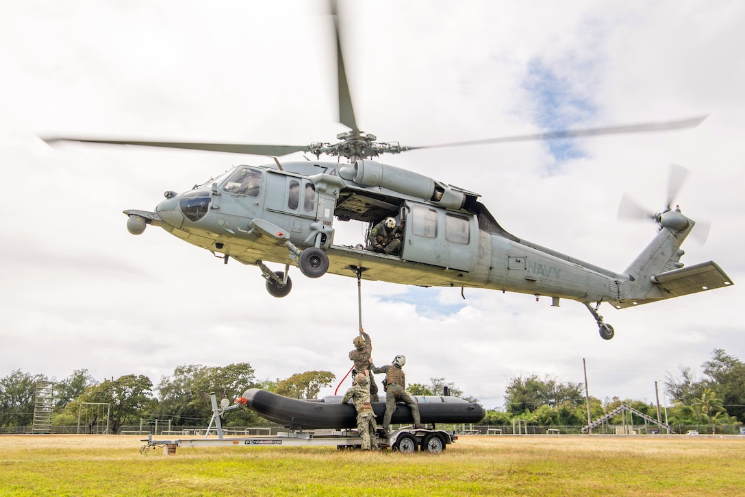 Sailors and Marines attach a rubber boat to an airborne helicopter in a field.