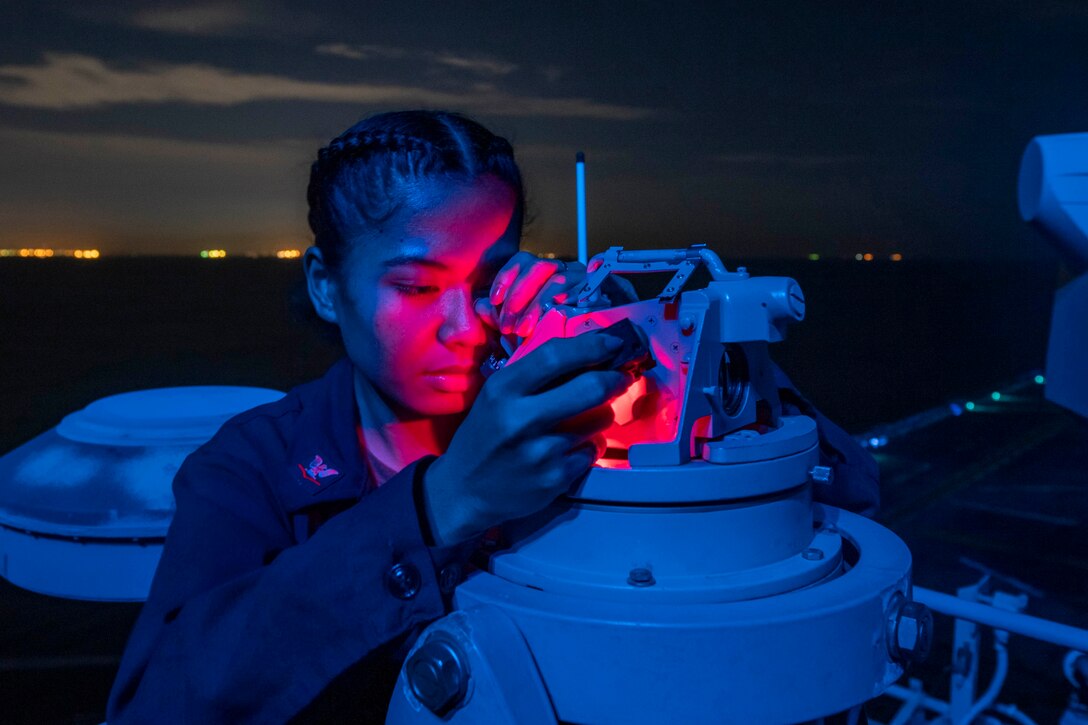 A sailor bathed in blue and red light checks an instrument on a ship at sea.