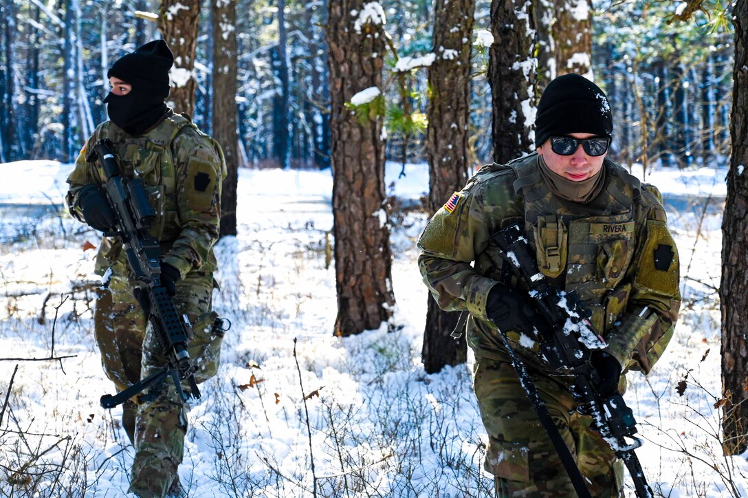 Two soldiers hold weapons while moving through a wooded area with snow on the ground.