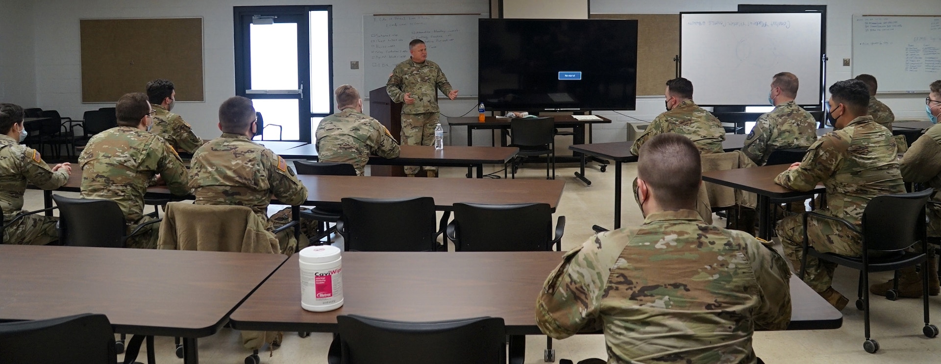 Around 20 people in Army OCP camouflage uniform sit at rectangular tables in a classroom.