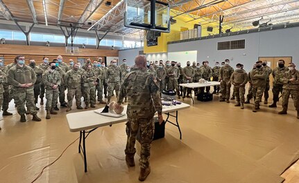 Around 40 people in Army OCP camouflage uniform stand in curved rows in a large gymnasium with natural light and light gold wood floors. They are looking at a tall man in uniform who is behind a table with a CPR  manakin.