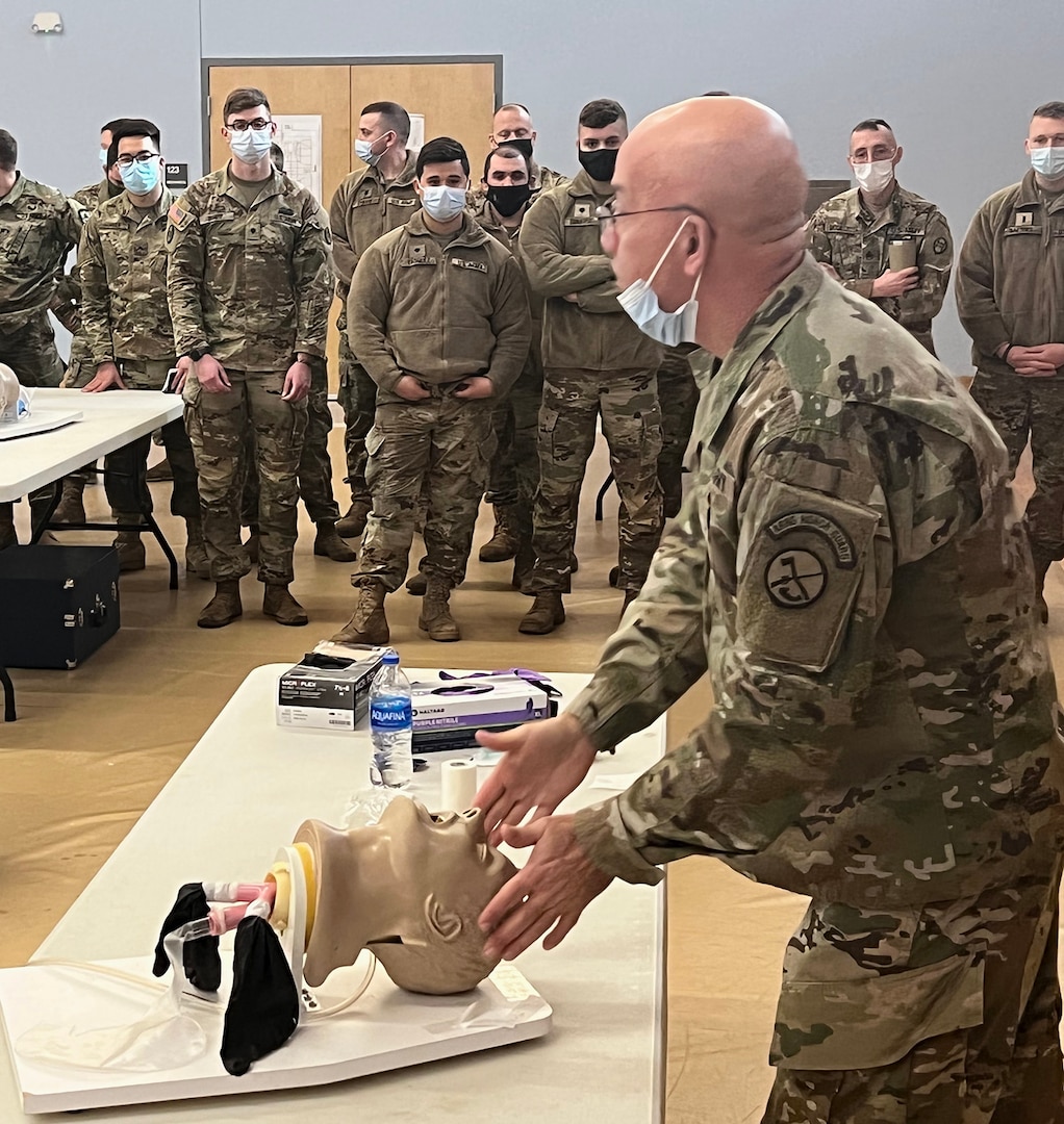 Looking from the left side: A tall man dressed in Army OCP camouflage uniform and wearing a face mask is holding a CPR manikins head that is displayed on a table. Around 20 Soldiers in uniform stand to the right of the man, relaxed, as they watch the demonstration.