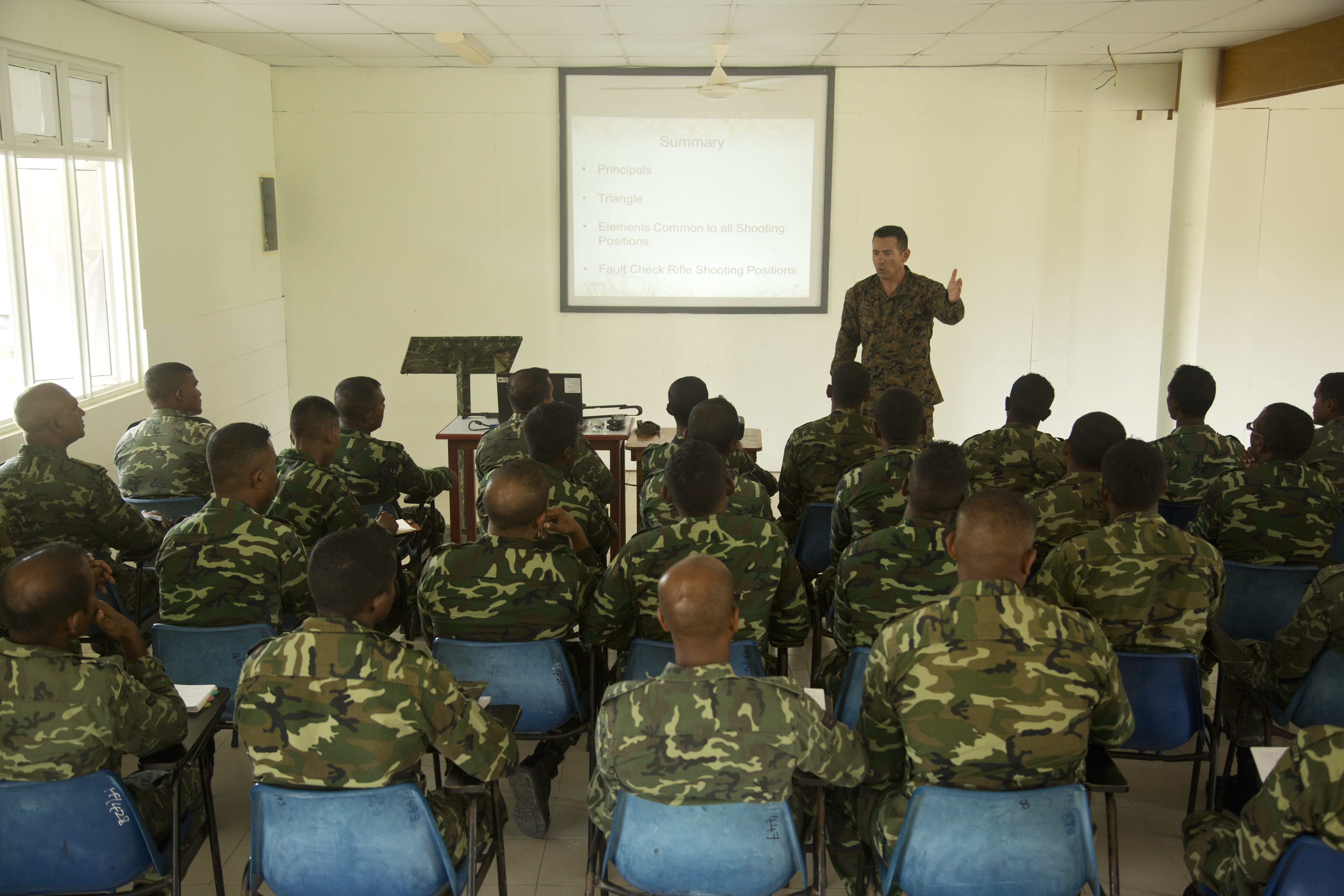 A Marine at the head of a classroom instructing more Marines sitting down with their backs to the camera.