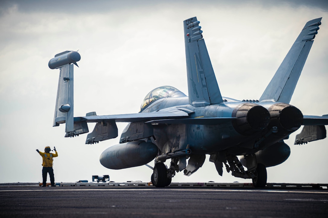 A sailor signals toward a jet on the deck of a ship.