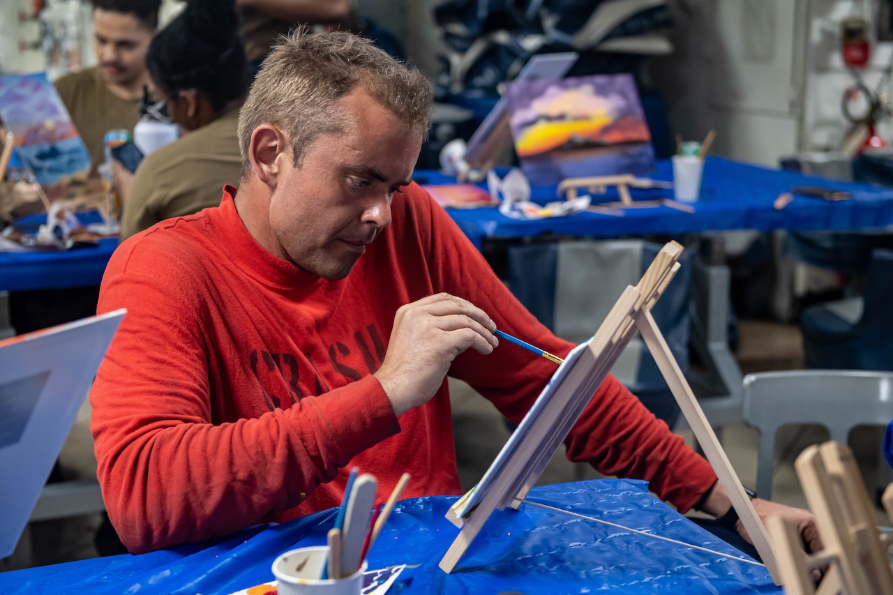 A sailor paints on a canvas while sitting near others doing the same.