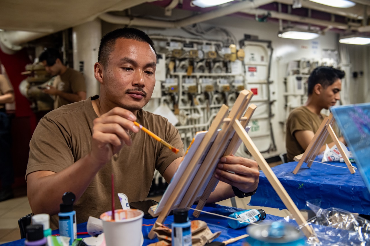 Two sailors sit at tables and paint using easels.