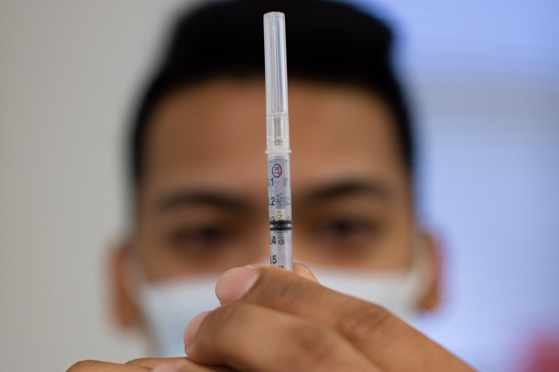 A corpsman wearing a face mask holds a syringe.