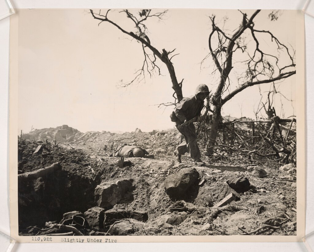 A Marine Rifleman crouches low to move forward as Japanese machine gun bullets rip through the trees above his head on Iwo To, Japan, during the Battle of Iwo Jima, Feb. 25, 1945. A Japanese soldier who had been killed by advancing Marines an hour earlier, lies on the ground.