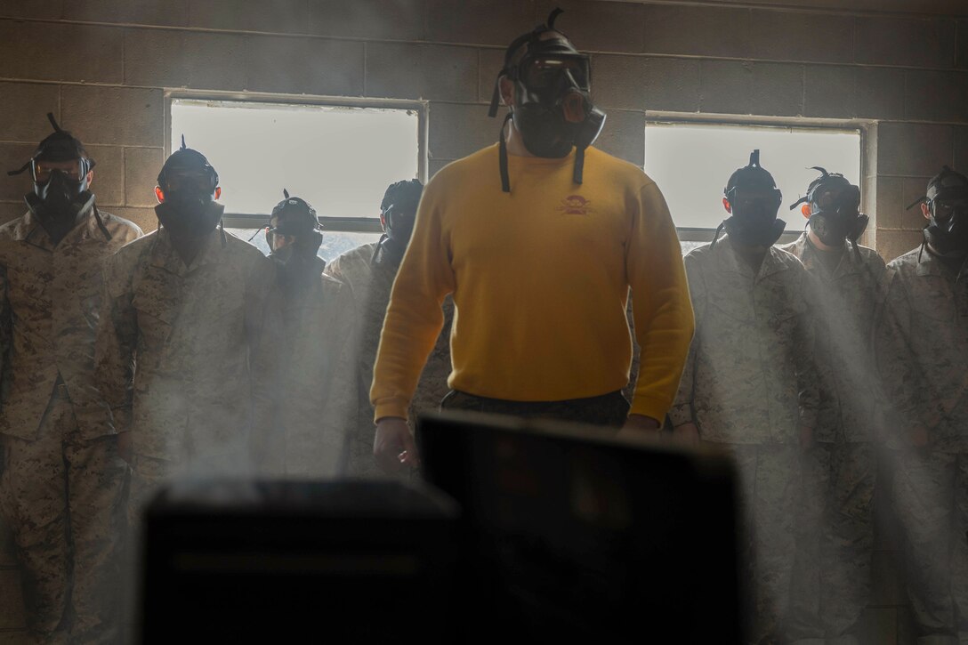 Marine Corps recruits wearing gas masks stand in a room surrounded by smoke during training.