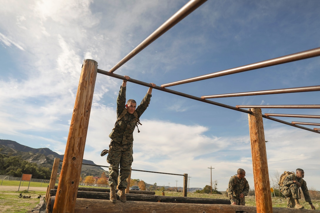 A Marine hangs on to a jungle gym as fellow Marines stand nearby.