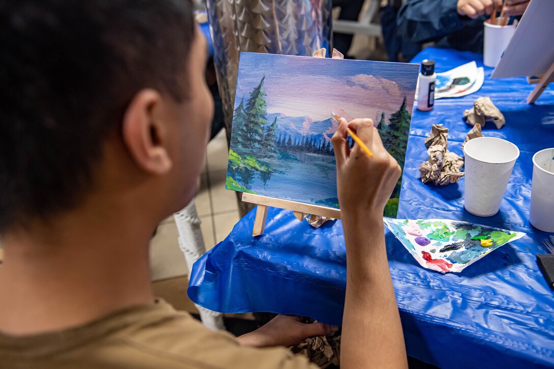 A sailor works on a painting while seated at a table with painting tools.