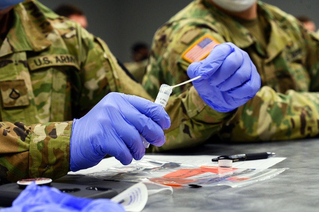 A soldier wearing gloves holds a plastic tube while inserting a nasal swab into it.