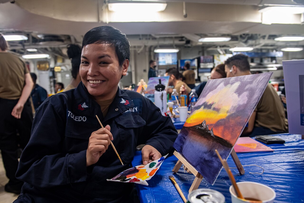 A sailor smiles and holds a paint brush while sitting next to a painting.