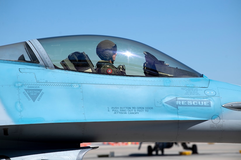 A pilot sits in the cockpit of a blue fighter jet.