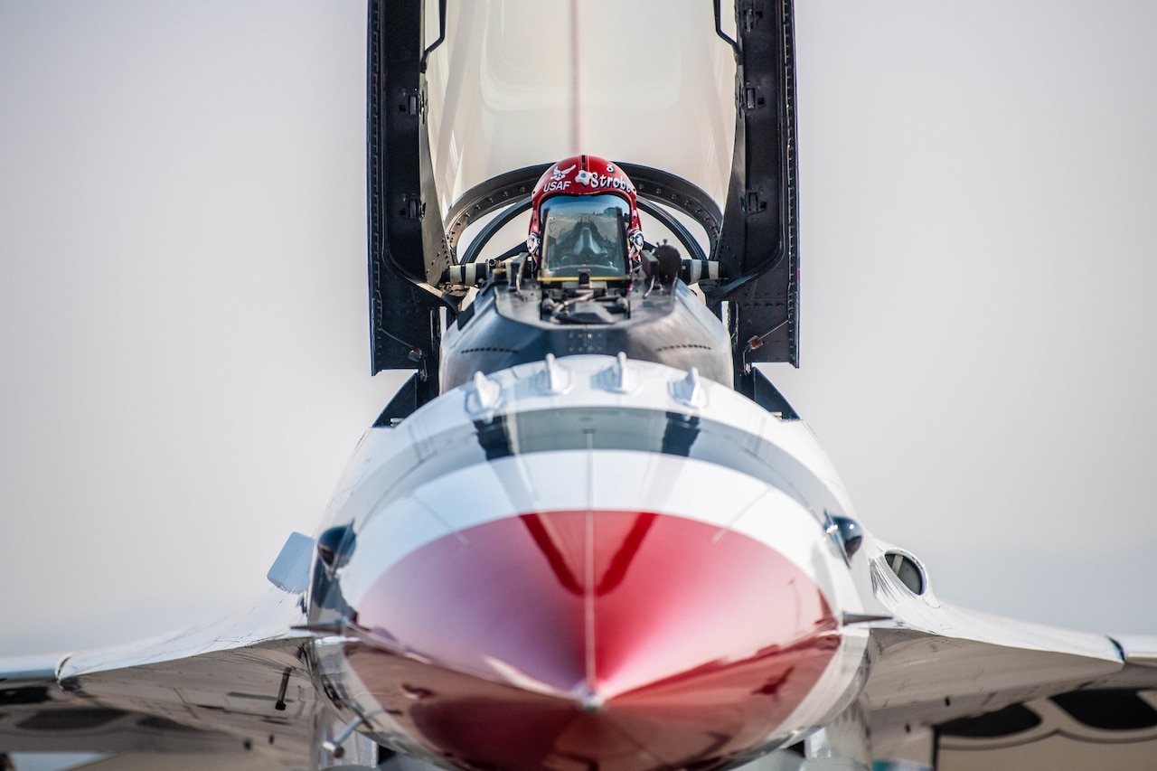 A helmeted pilot sits in a fighter jet with the canopy open.