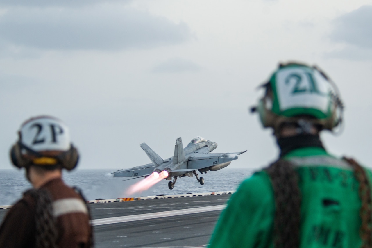 A fighter jet takes off from an aircraft carrier deck. Two people watch in the foreground.