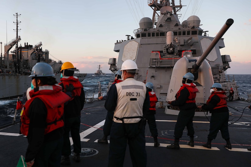 Sailors stand on deck looking toward another ship that has pulled up alongside.