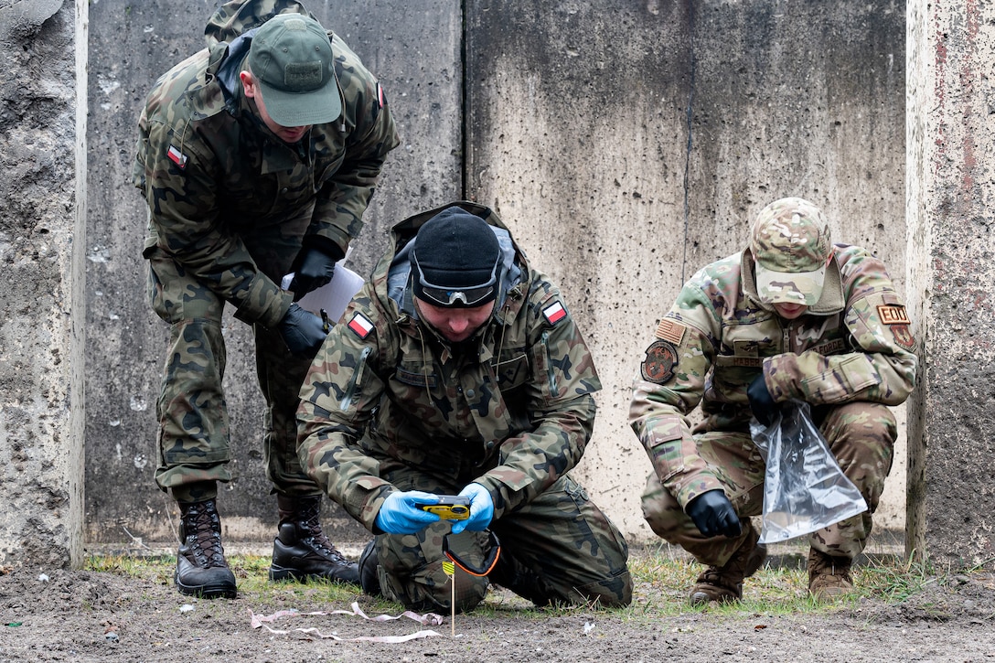 Three men look at the ground as they gather pieces of shrapnel.