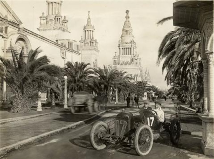 A race car driver cruises down a tree-lined street.