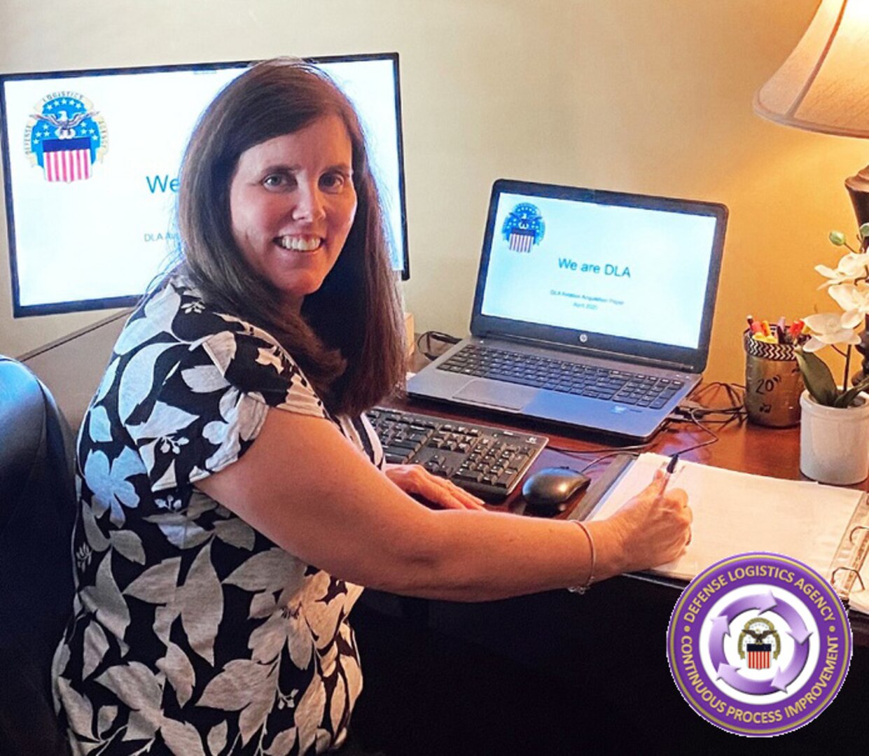 A woman sits at a desk posing for a photo in front of monitors displaying the logo for the Defense Logistics Agency and "We are DLA." At the bottom left is a logo for DLA Troop Support Office of Continuous Process Improvement.