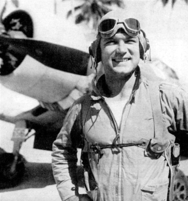 A man with flight goggles on his forehead stands in front of a propeller airplane.