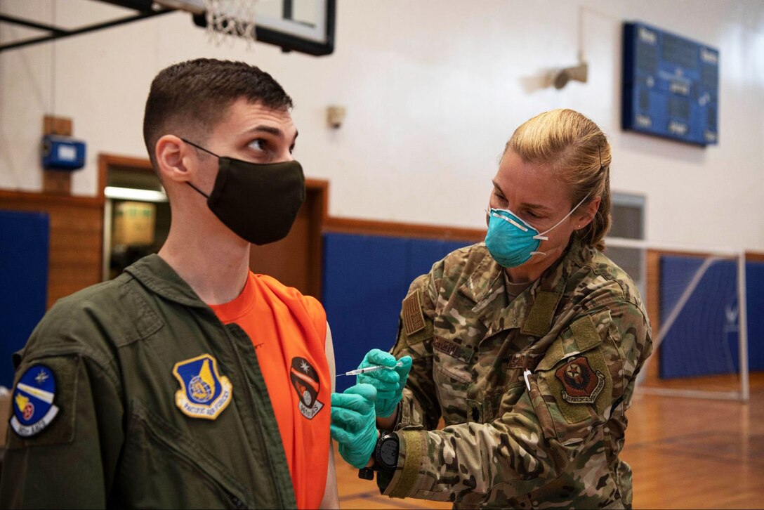 An airman wearing a face mask and gloves vaccinates another airman wearing a face mask.