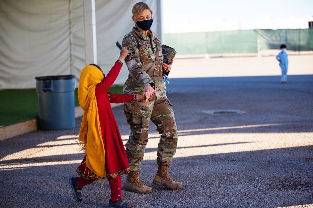 An Air Force officer wearing a face mask walks alongside an Afghan child.