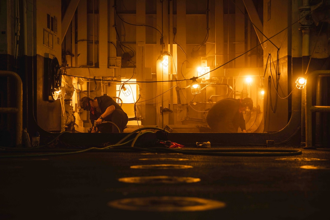 Two sailors work on a ship's deck at night with bright lights around them.