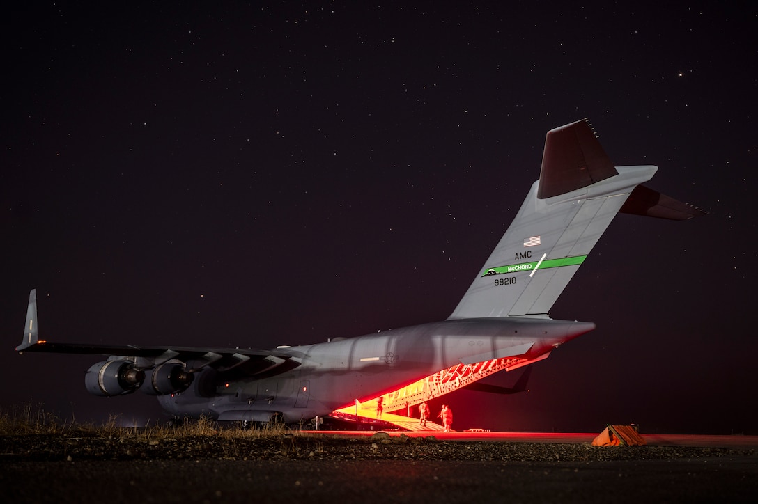 Airmen and soldiers walk on an aircraft’s ramp under a starry sky illuminated by  an orange light.