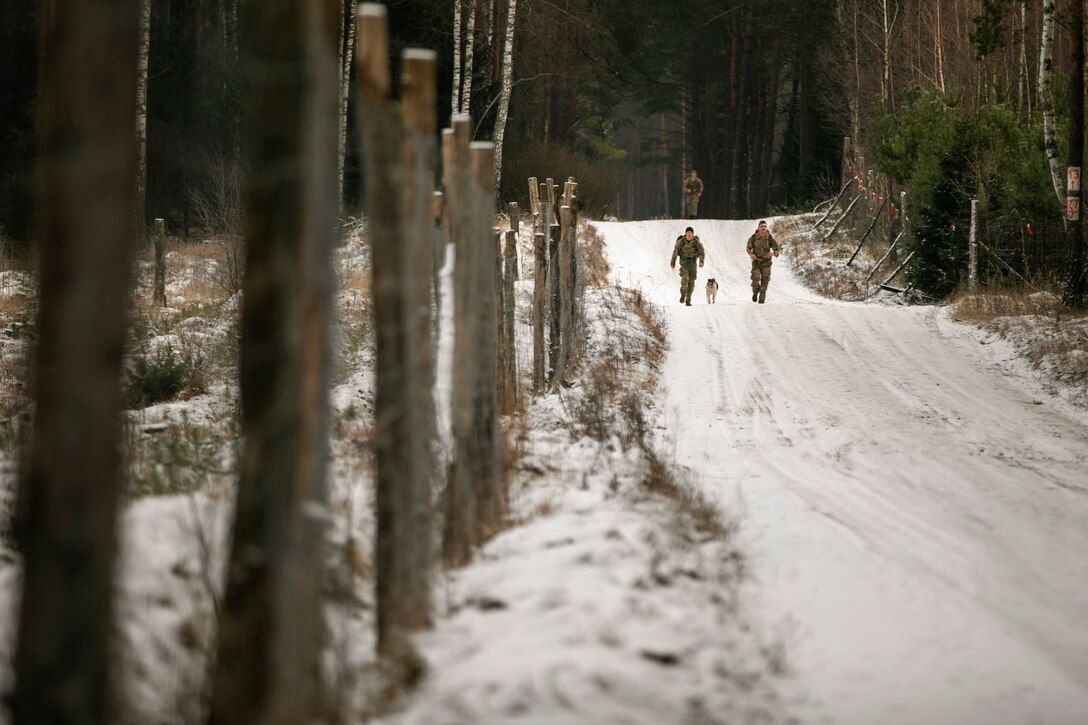 Guardsmen walk with a dog on a snowy path in the woods.