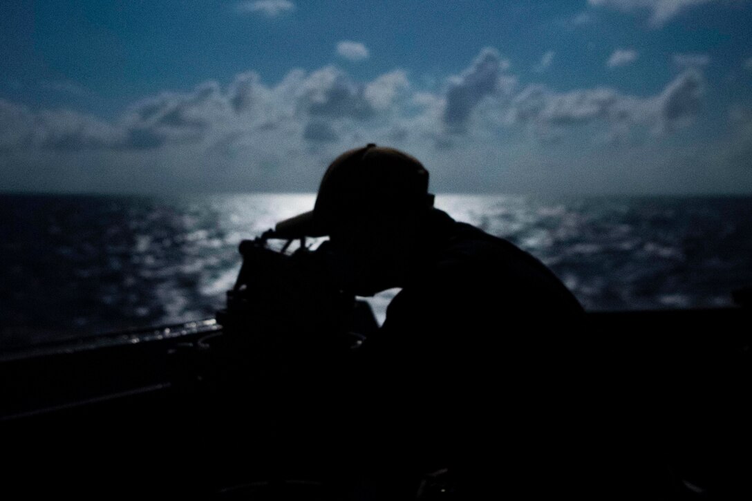 A sailor shown in silhouette stands watch on a ship.