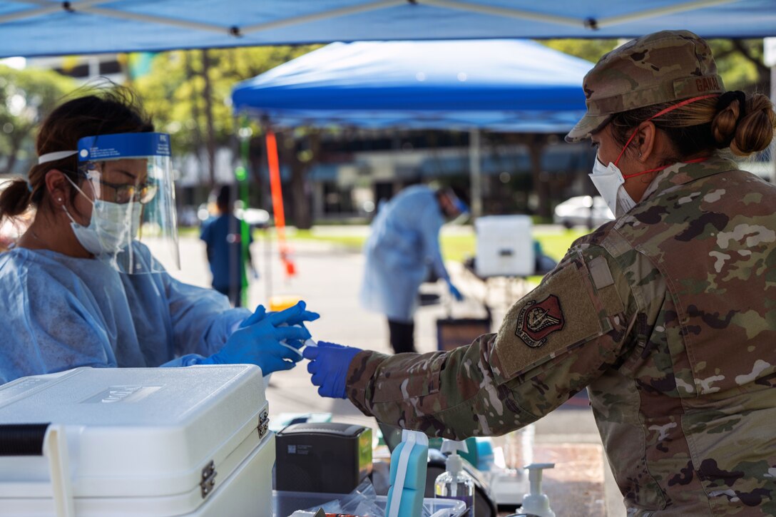 A soldier, wearing a face mask and gloves, gives an unopened tube to a medical tech wearing personal protective equipment.