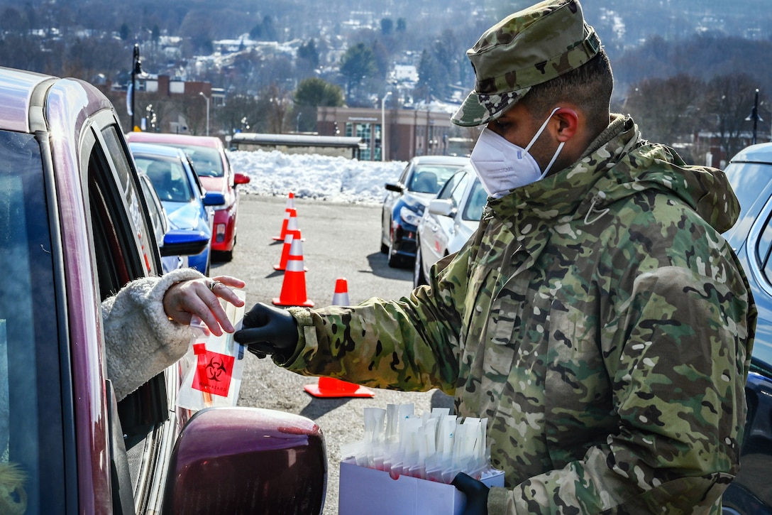 A soldier wearing a face mask and gloves hands a COVID-19 test kit to a person through the window of a car.