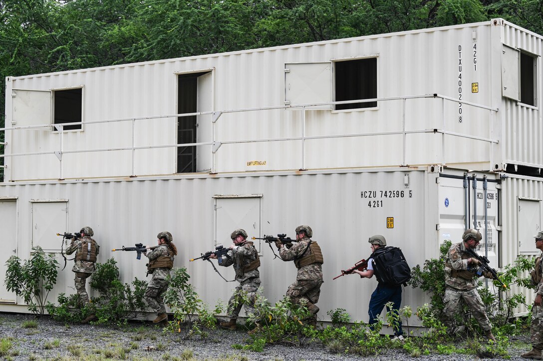 Airmen holding weapons move in a line around a building.