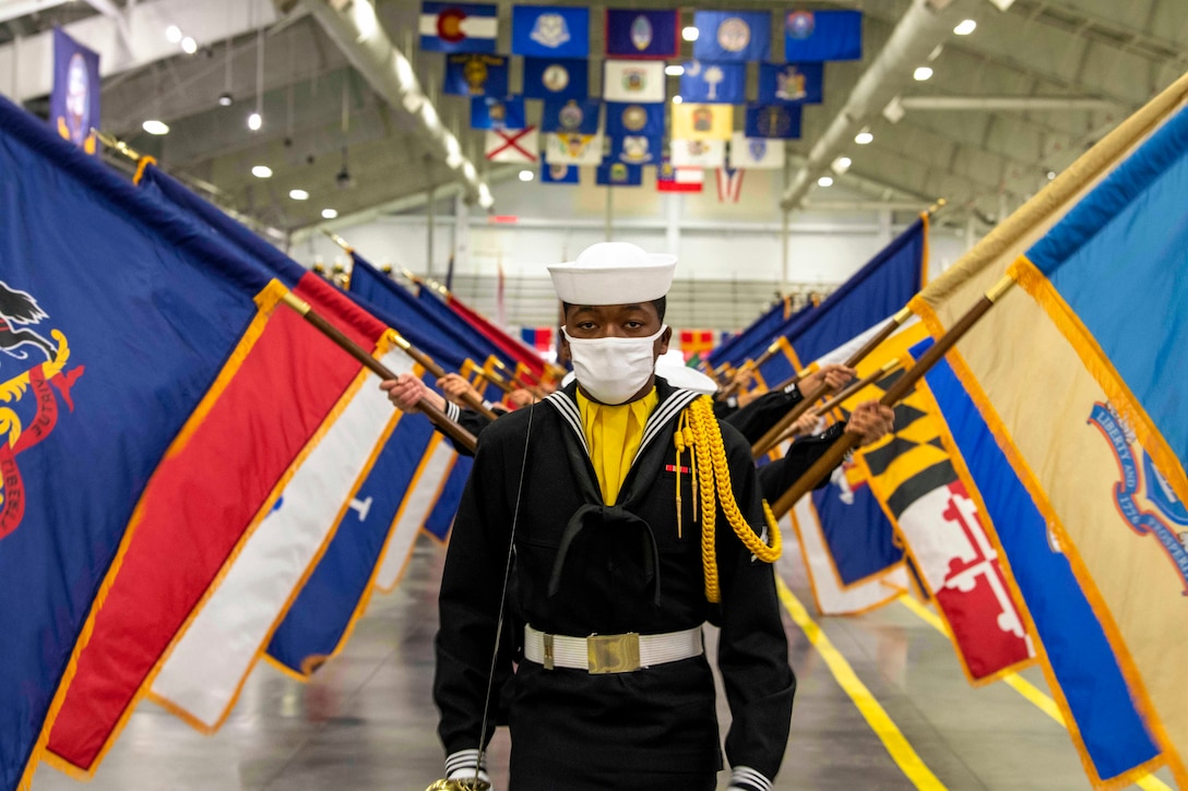 Sailors wearing face masks stand in a line holding flags.