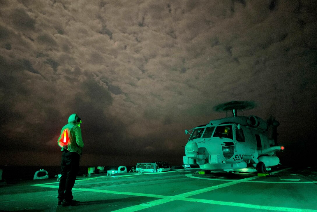 A sailor holding a bright light stands in front of a helicopter aboard a ship under a cloudy sky.