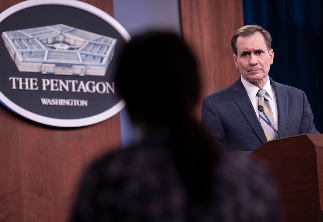 A man stands at a lectern and addresses a person who is in silhouette. The sign on the wall indicates that they are at the Pentagon.