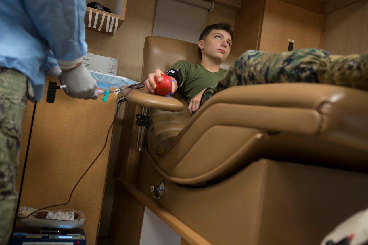 A service member holds a stress ball while lying on a hospital bed as another person in uniform prepares to draw his blood.