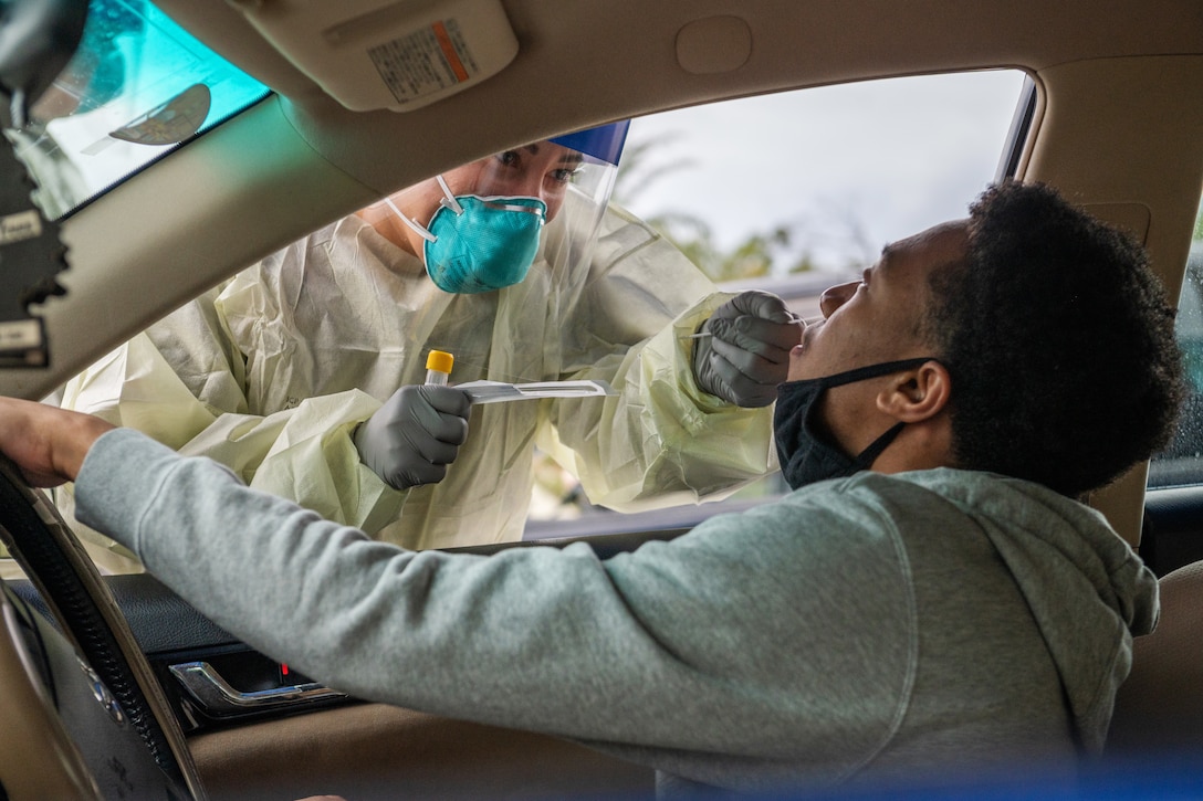 An airman wearing personal protective equipment holds a nasal swab while administering a COVID-19 test to man seated in a car.