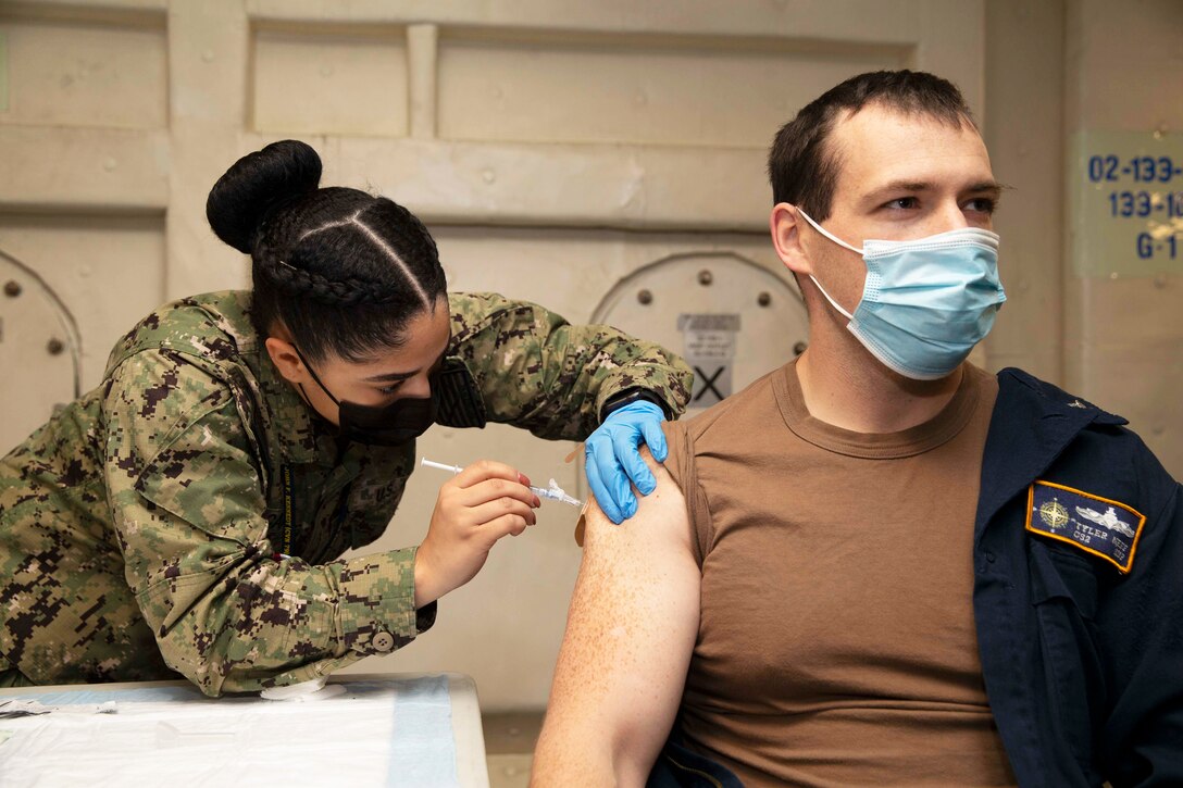 A Navy seaman wearing a face mask and gloves gives a COVID-19 booster vaccine to another sailor.