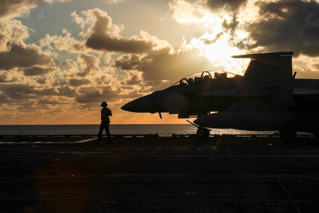 A sailor stands next to a large jet on the deck of a military ship.