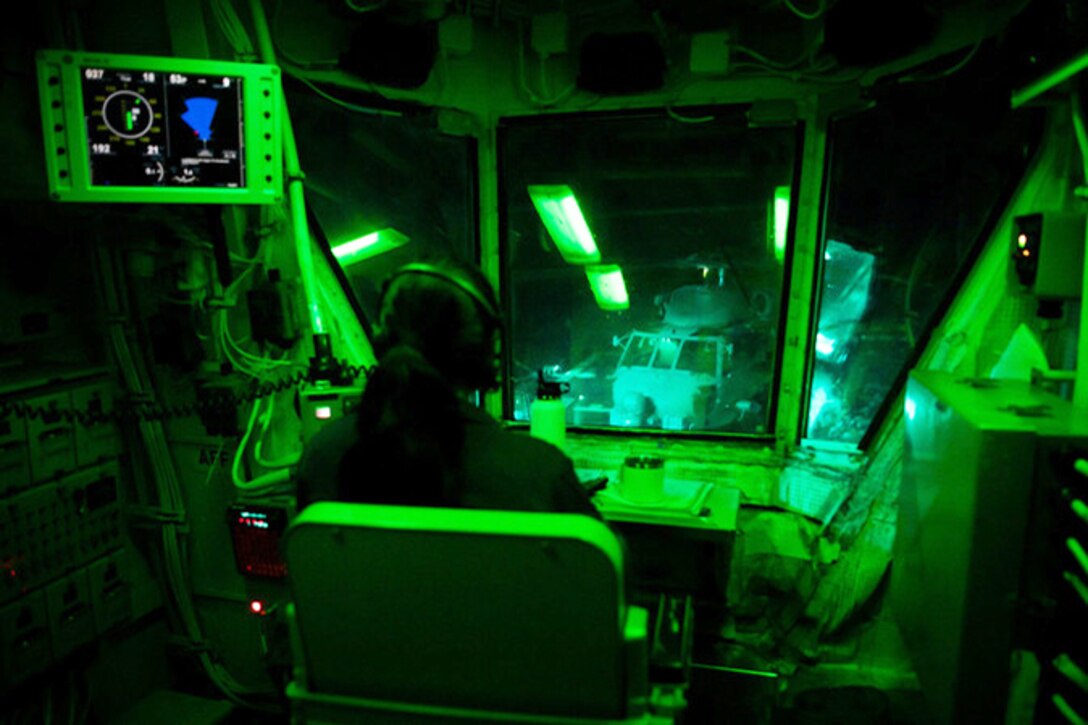 A sailor sits in a room lit with green light looking out large windows.