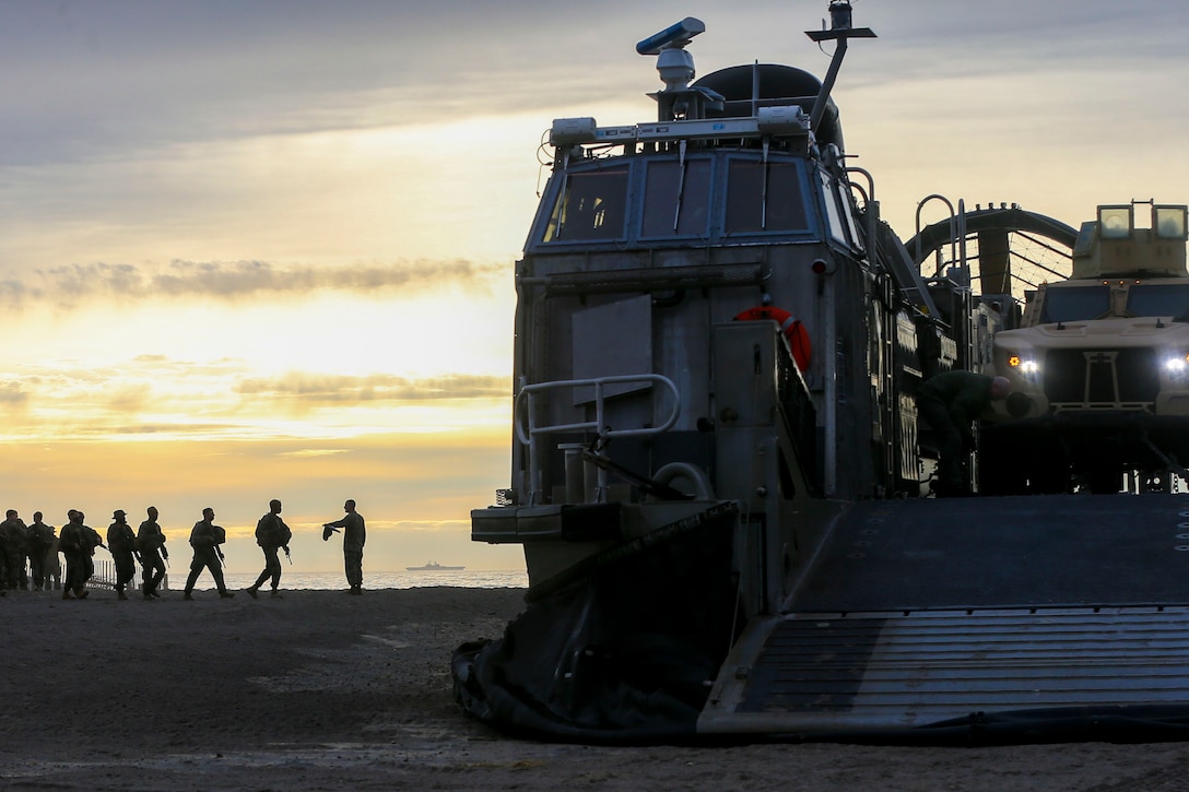 Marines walk on a beach toward an air-cushioned landing craft.