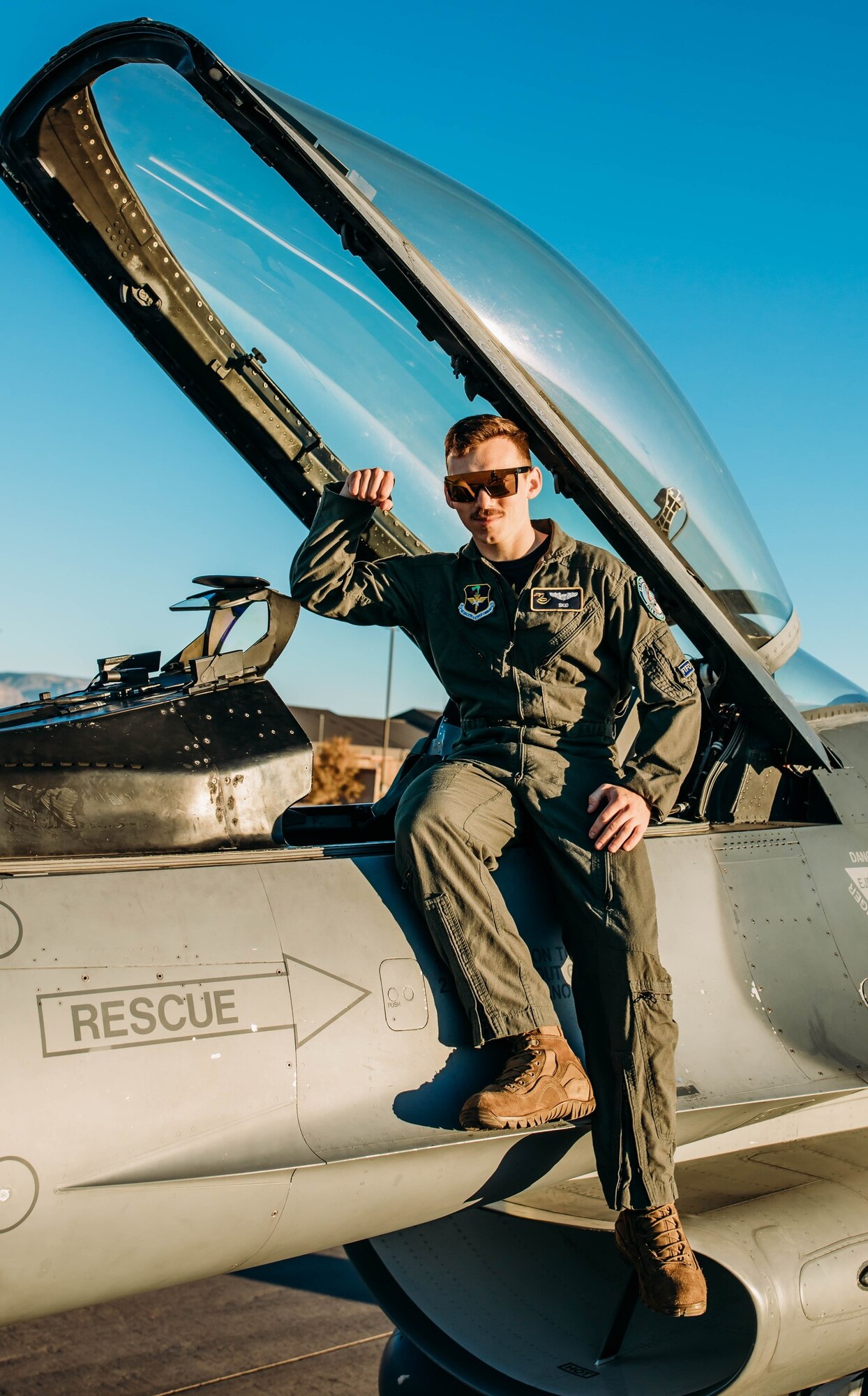 A pilot in a green flight uniform holds their arm up while sitting on an F-16 Fighting Falcon in front of a blue sky.
