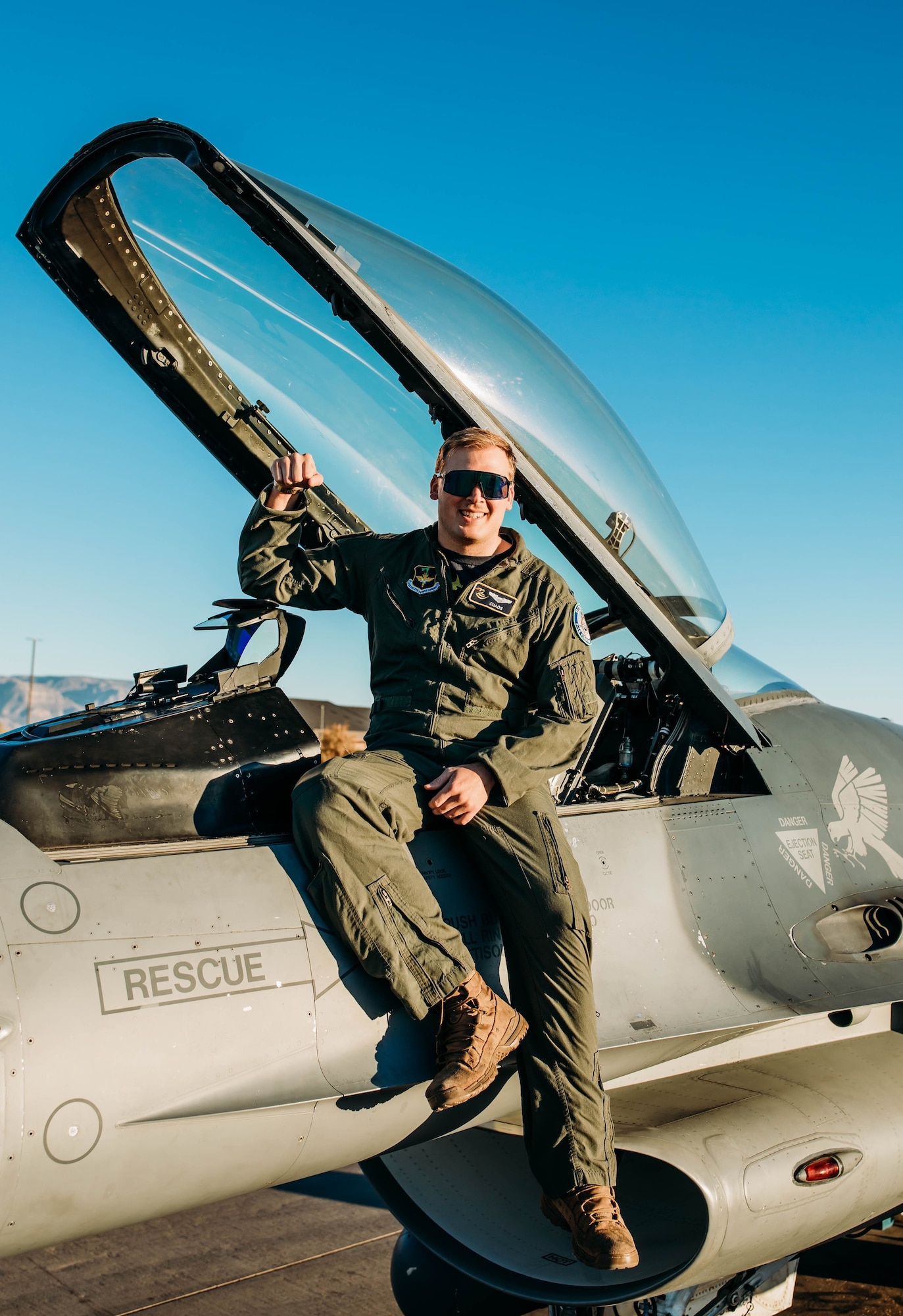 A pilot in a green flight uniform holds their arm up while sitting on an F-16 Fighting Falcon in front of a blue sky.