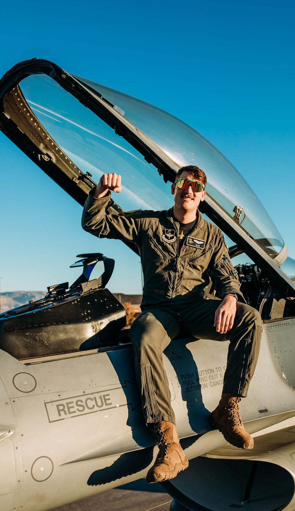 A pilot in a green flight uniform holds their arm up while sitting on an F-16 Fighting Falcon in front of a blue sky.