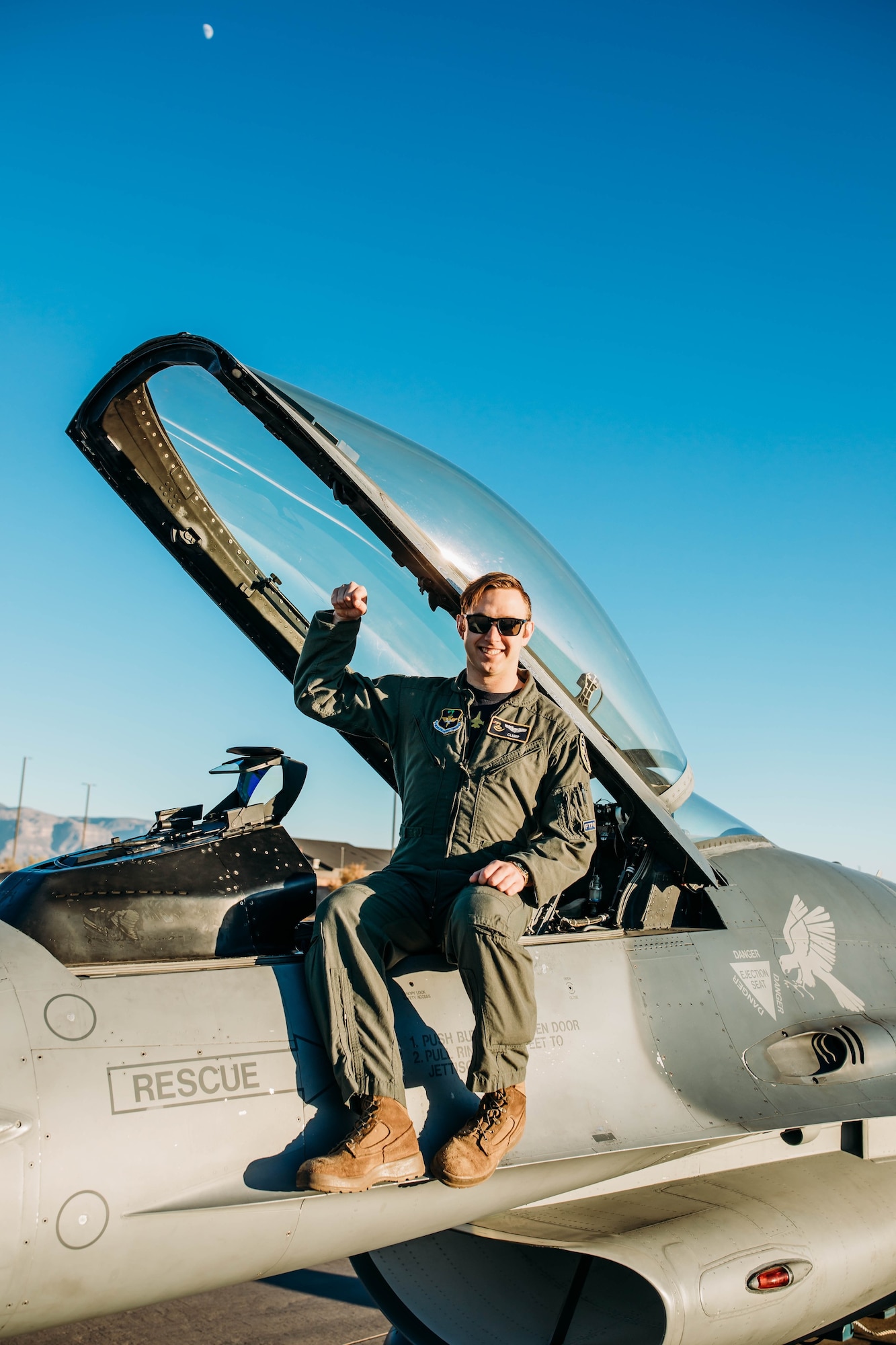 A pilot in a green flight uniform holds their arm up while sitting on an F-16 Fighting Falcon in front of a blue sky.