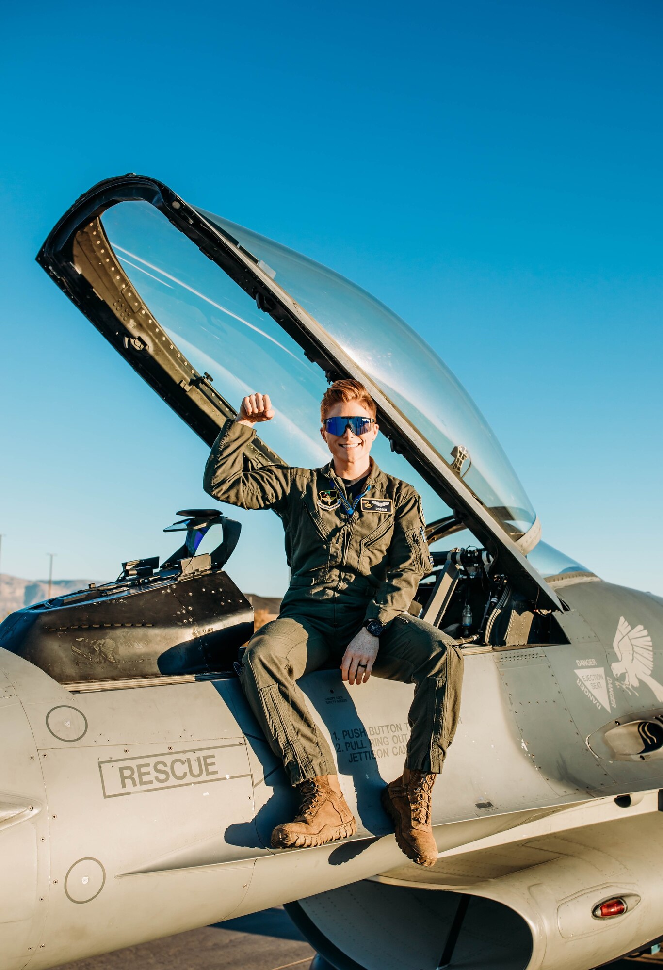 A pilot in a green flight uniform holds their arm up while sitting on an F-16 Fighting Falcon in front of a blue sky.