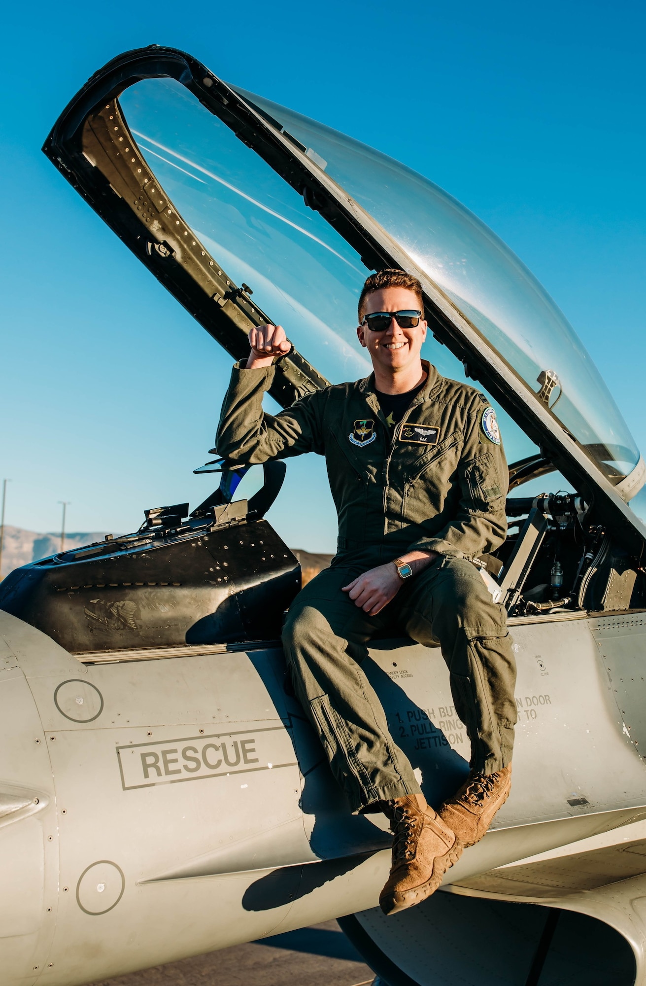 A pilot in a green flight uniform holds their arm up while sitting on an F-16 Fighting Falcon in front of a blue sky.