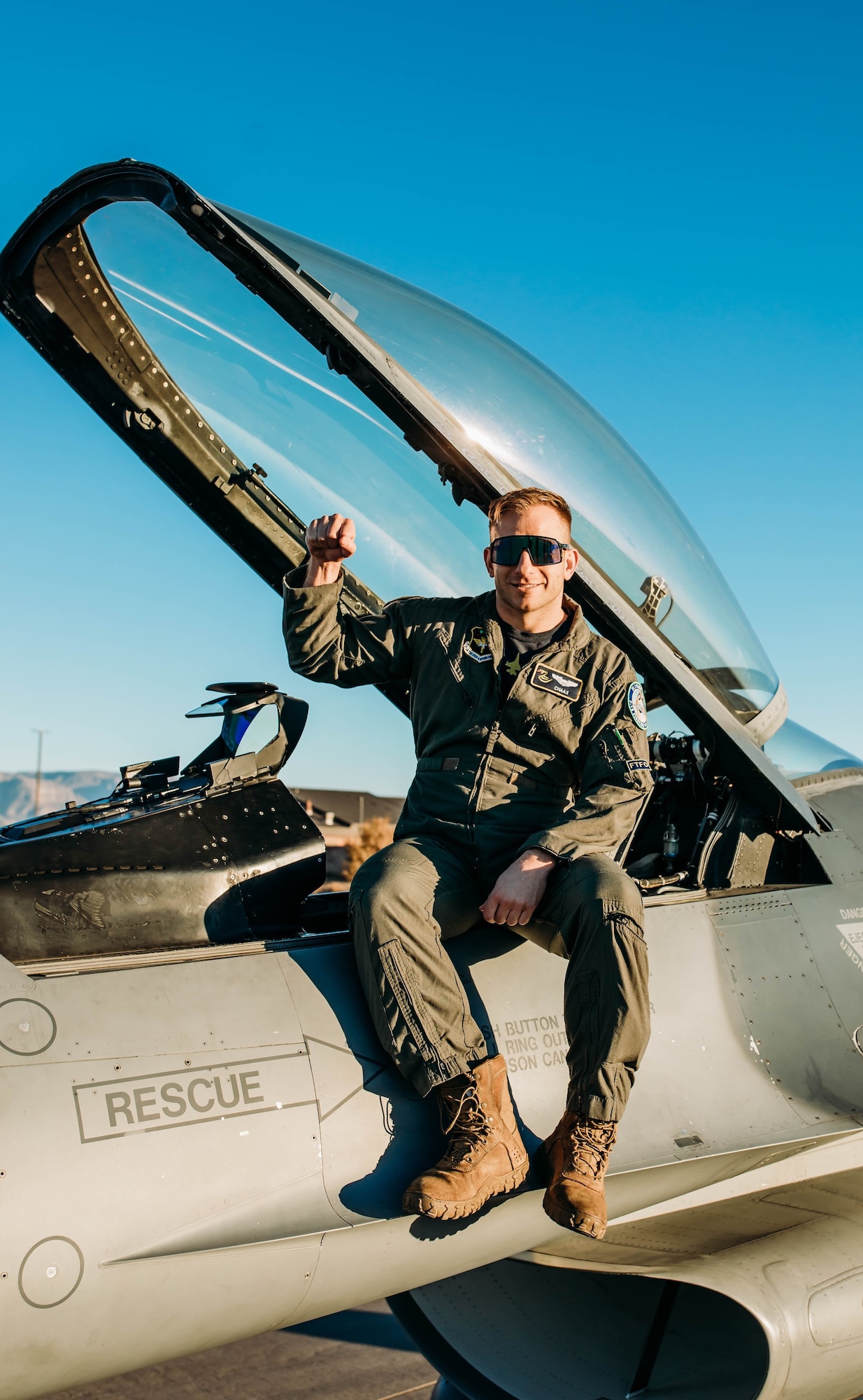 A pilot in a green flight uniform holds their arm up while sitting on an F-16 Fighting Falcon in front of a blue sky.